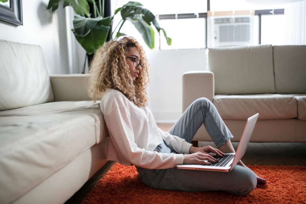 Uma mulher está sentada no chão de seu apartamento, conferindo as dicas para morar sozinho em seu notebook.