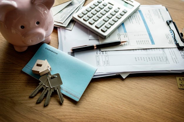 Na imagem temos um cofrinho, tabelas de gastos da casa e uma chave, destacando a importância do controle financeiro quando se está morando sozinho.